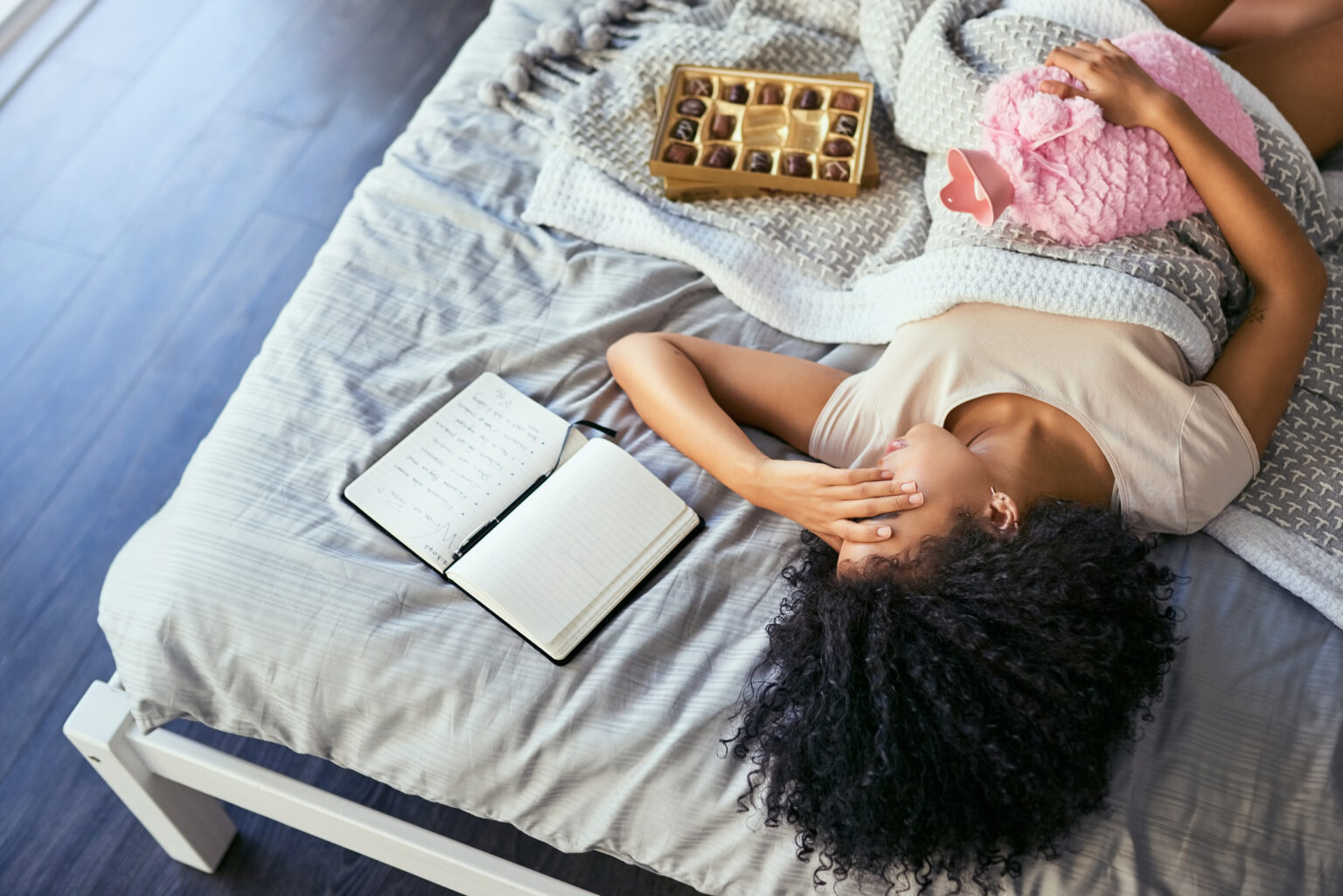 Young woman lying on a bed with a hot water bottle on her abdomen, appearing to be in menstrual pain, surrounded by a journal and chocolates — symbolizing the emotional and physical toll of hormonal imbalances potentially worsened by climate change and environmental stressors.